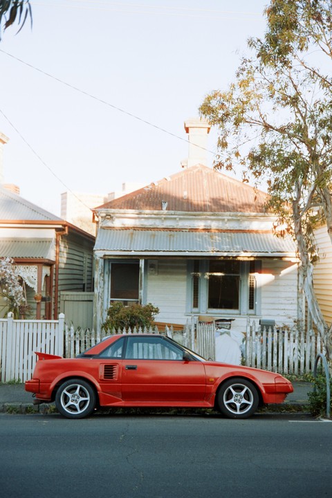 A red car is parked on the street in front of a house with a white picket fence.