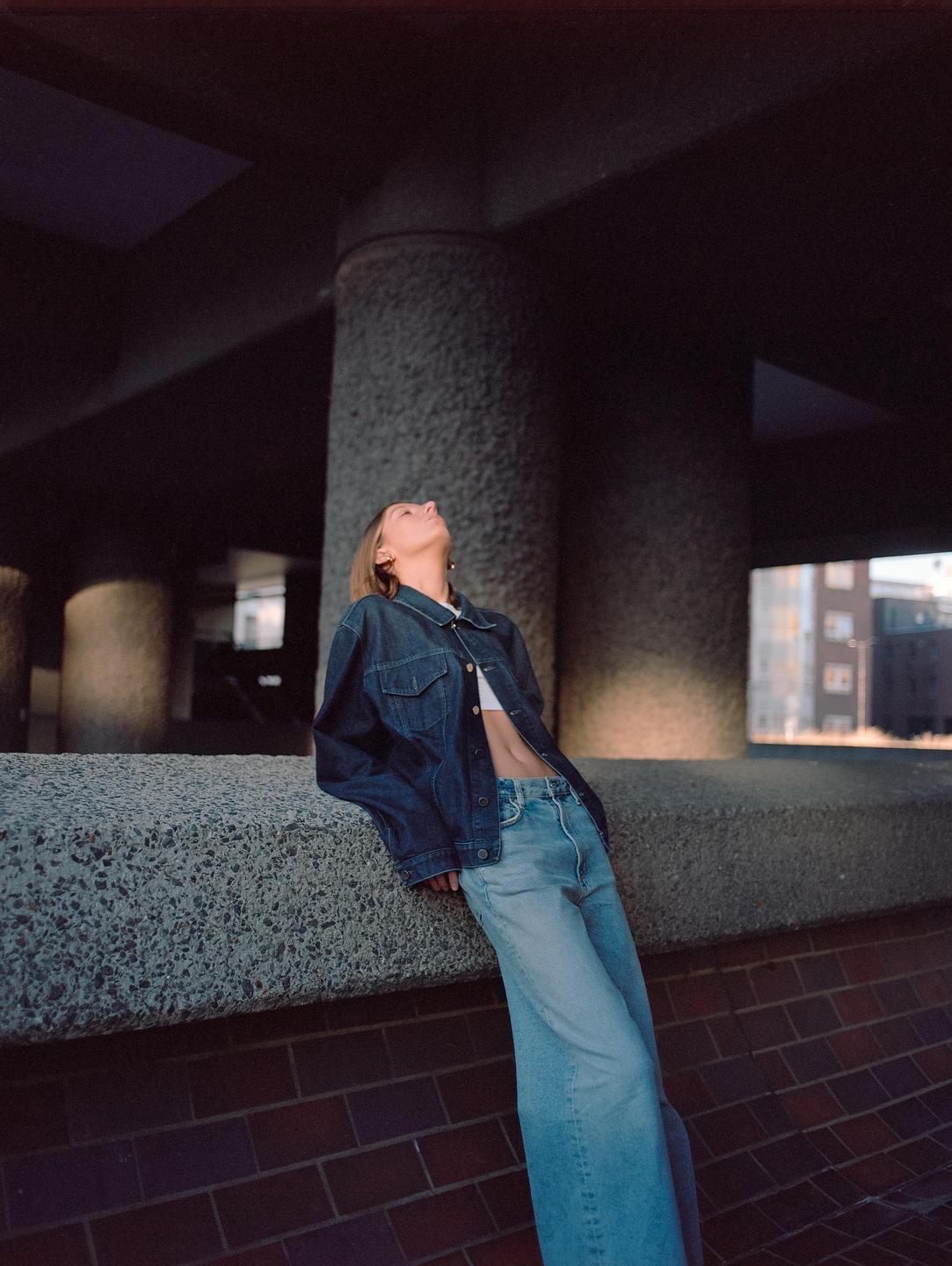 A woman in a denim jacket and jeans leans against a concrete wall and looks up.