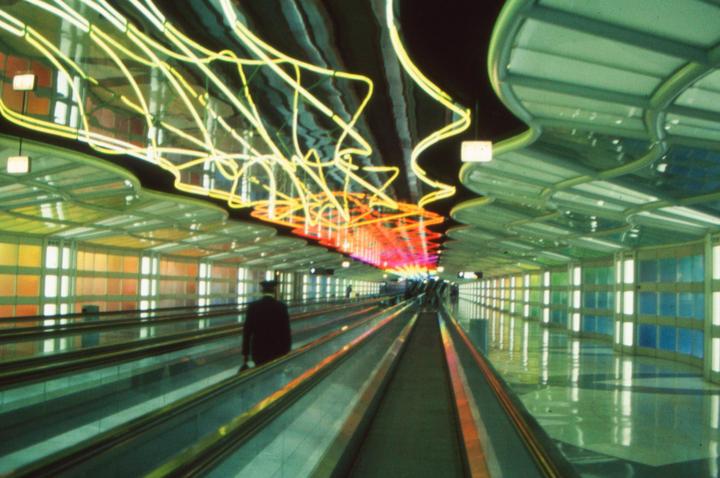 A man stands on a moving walkway in an airport tunnel with colorful lights and neon art.