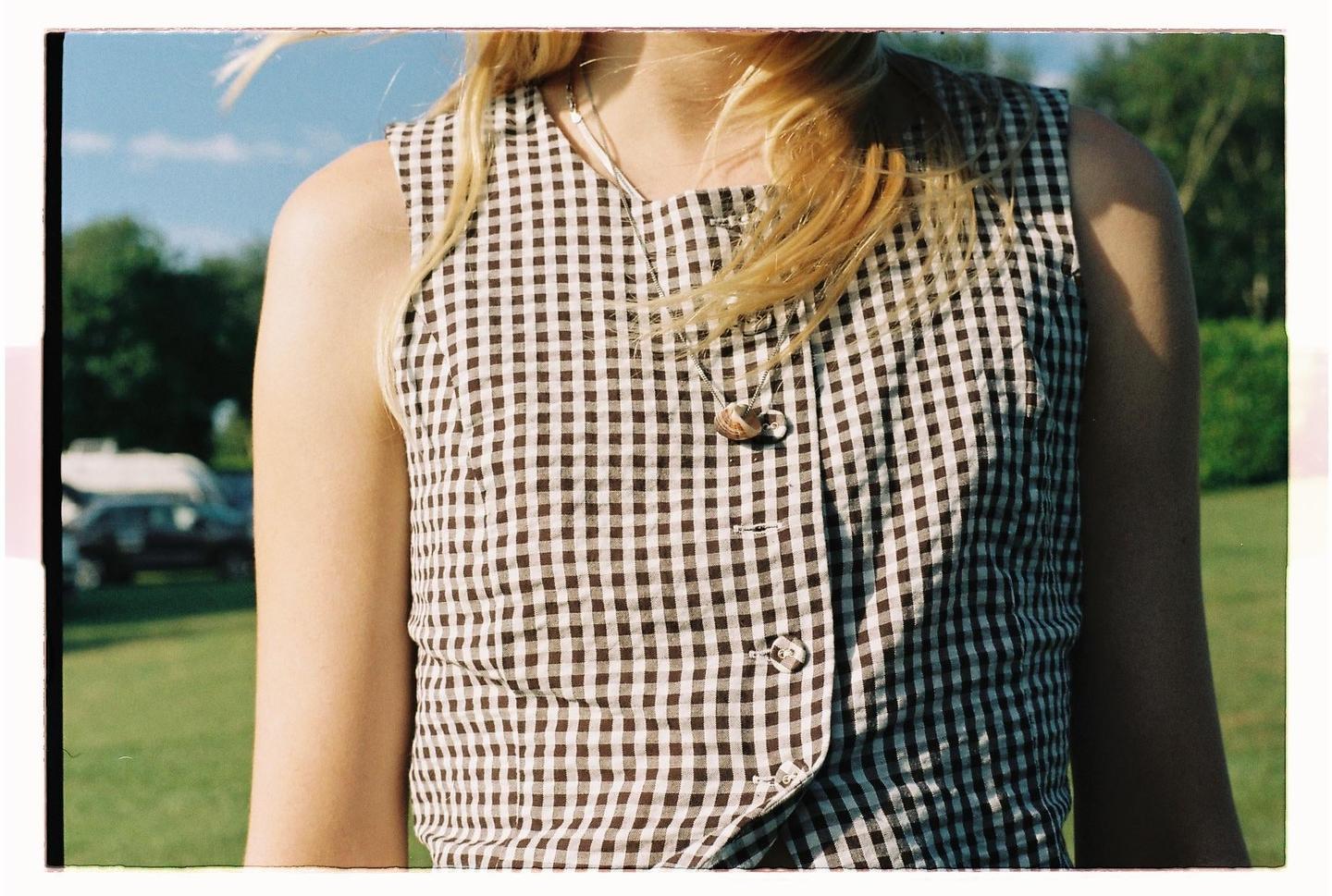 A woman wearing a checkered top and a necklace stands in a grassy field under a blue sky.
