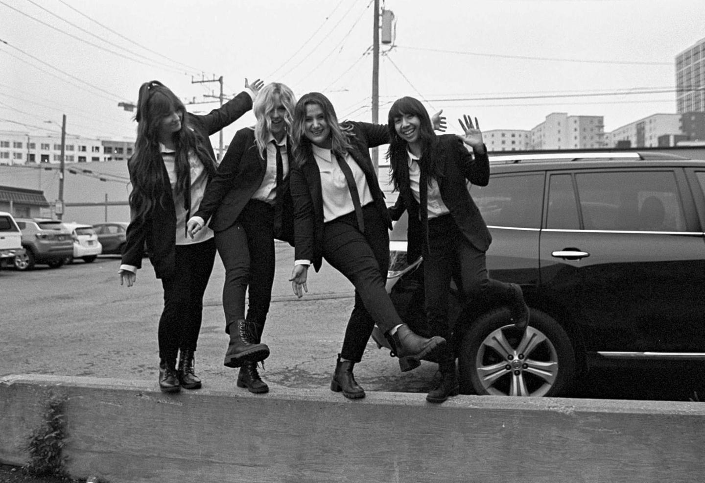 Four women in suits and ties pose playfully on a concrete barrier next to a parked car.