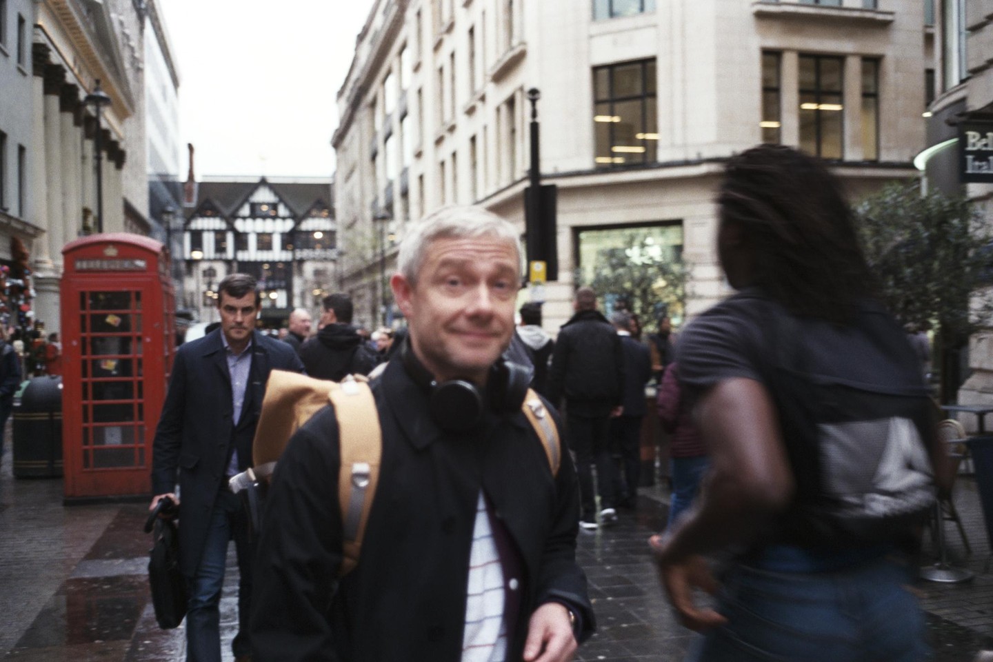 A man with headphones around his neck smiles at the camera while walking on a city street with a red telephone booth and other people.