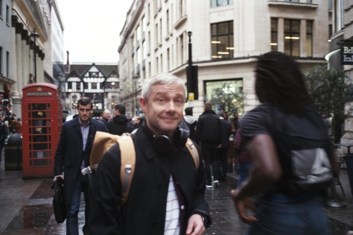 A man with headphones around his neck smiles at the camera while walking on a city street with a red telephone booth and other people.