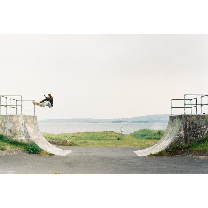 A man on a skateboard performs an aerial trick over a concrete ramp next to a body of water.