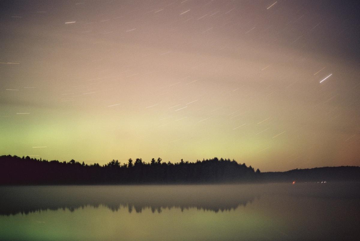 Star trails are visible above a forest reflecting in a misty lake at night.