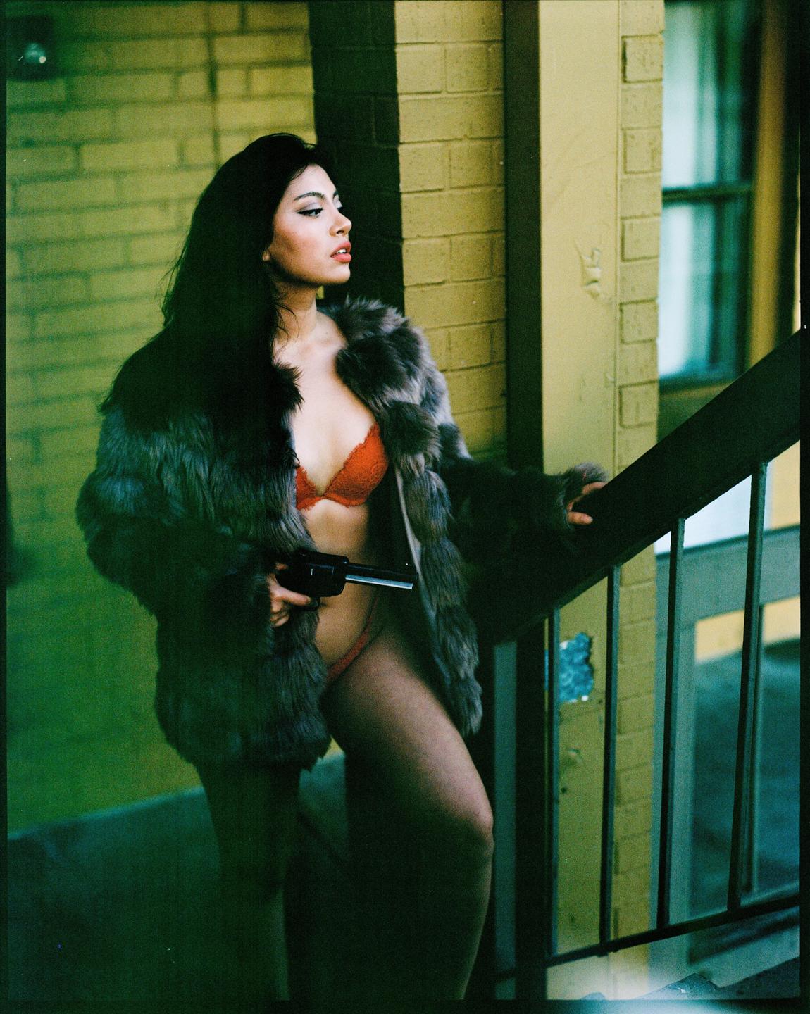 A woman in a fur coat and red bra holds a gun while standing on a staircase.