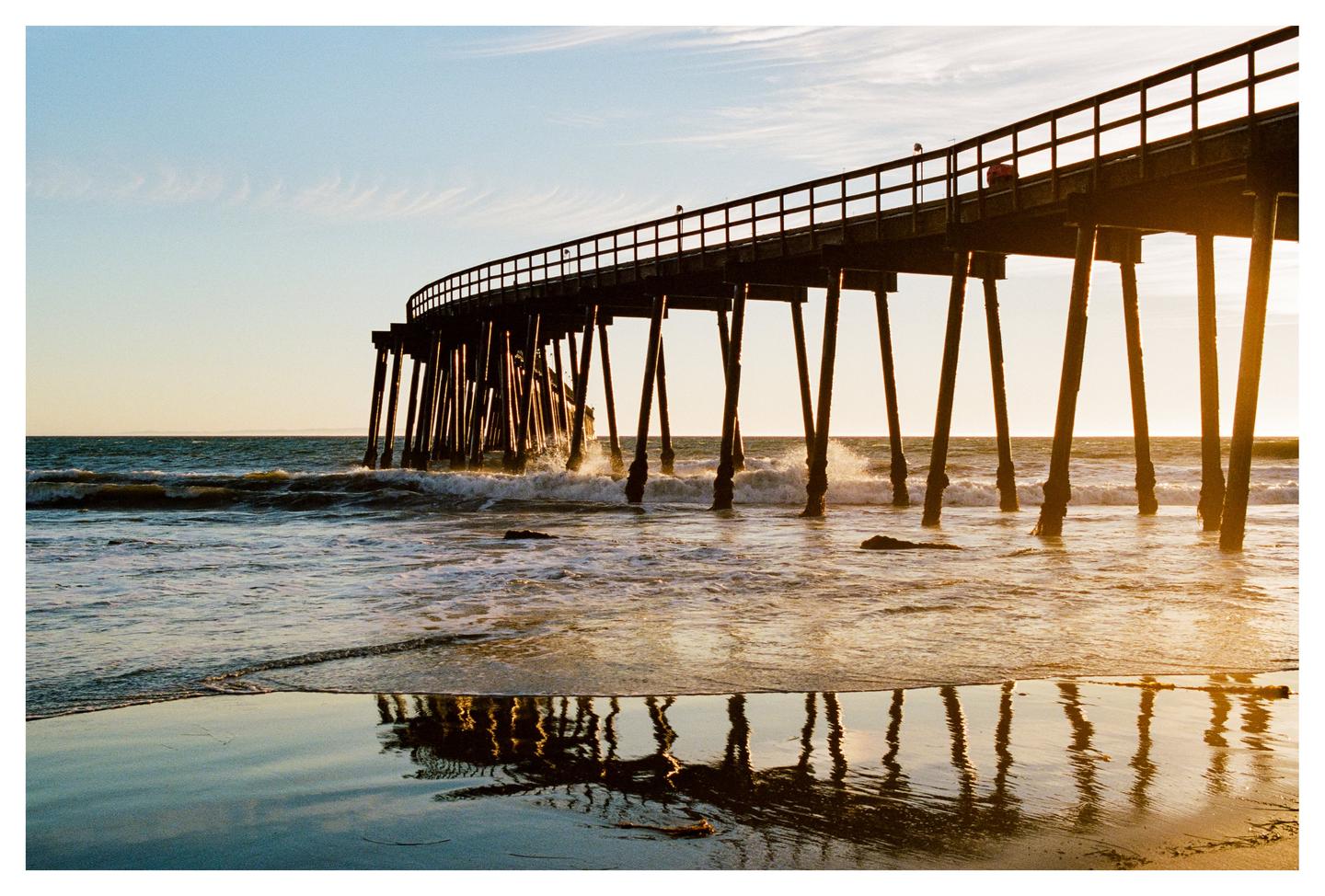 A long pier curves over the ocean with waves crashing against its supports at sunset.