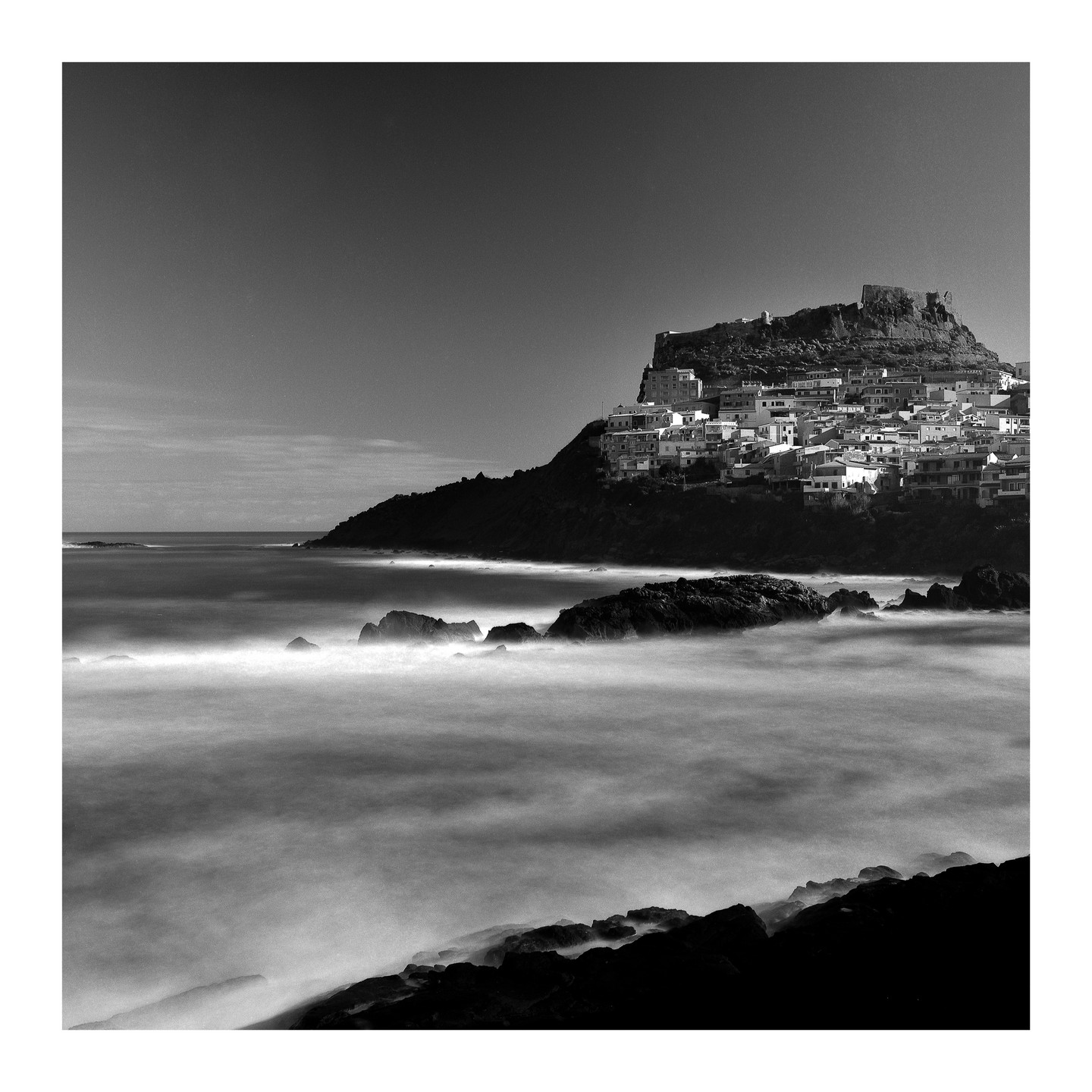 A village with white buildings is perched on a hill overlooking a rocky coast with blurred water.