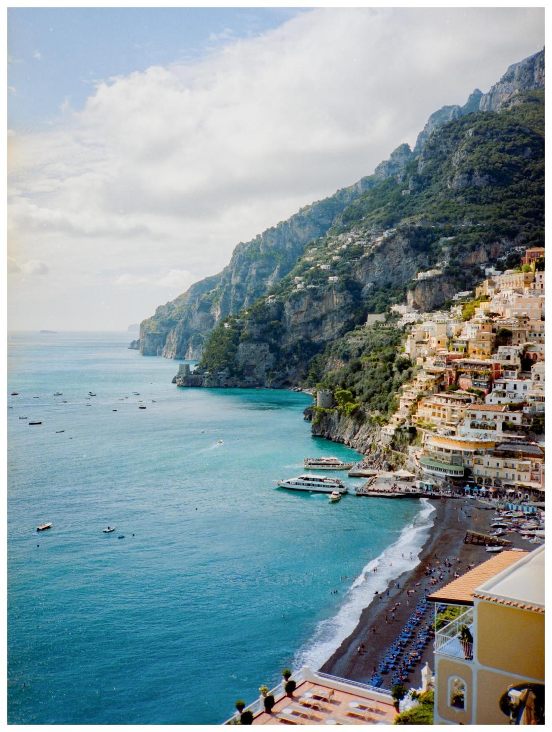 A coastal town with colorful buildings is nestled into a steep hillside overlooking a bay with boats and a beach.