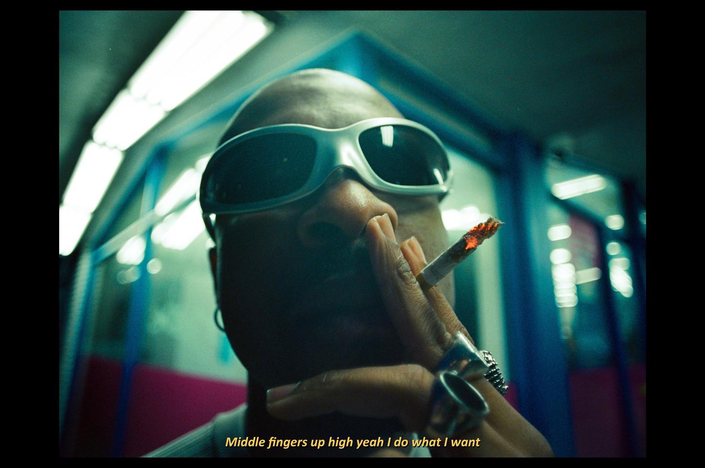 A man wearing sunglasses and rings holds a lit cigarette.