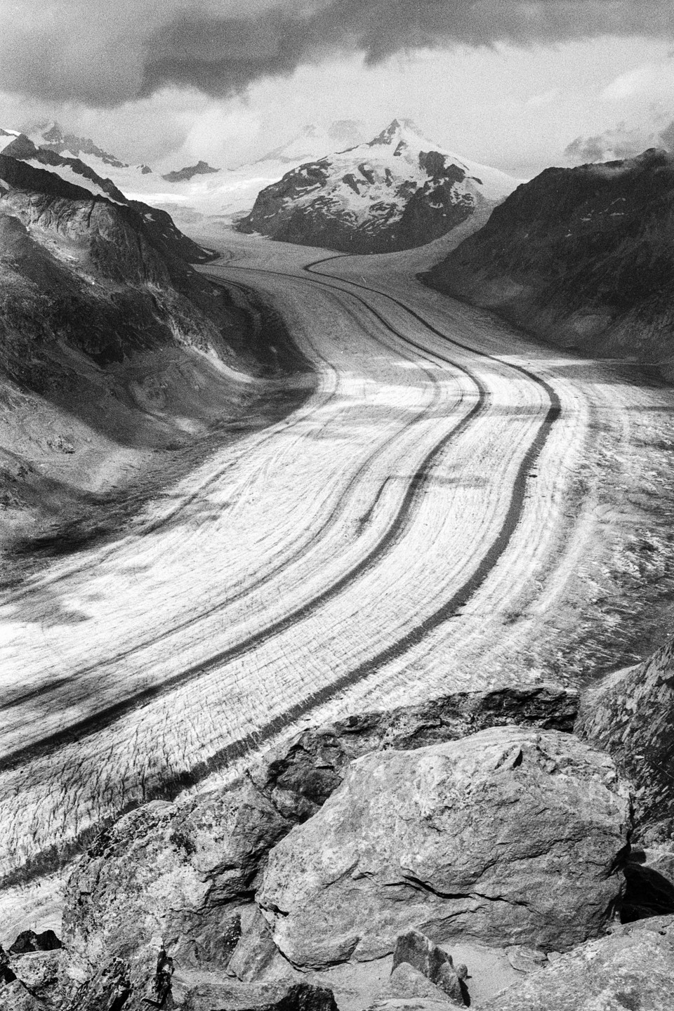 A wide glacier winds through a valley between snow-capped mountains under a cloudy sky.
