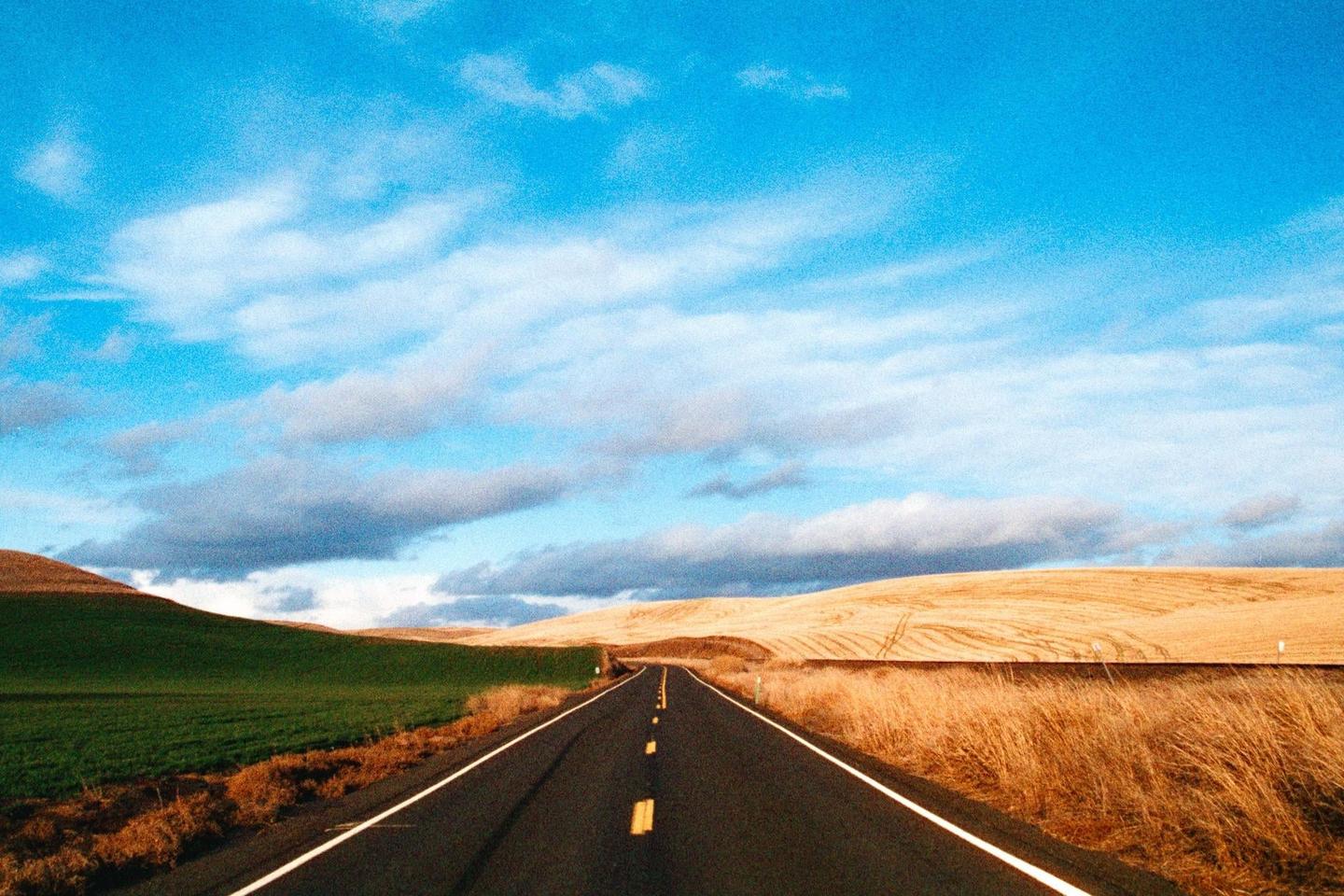 A long road stretches through rolling hills with green and golden fields under a blue sky with clouds.
