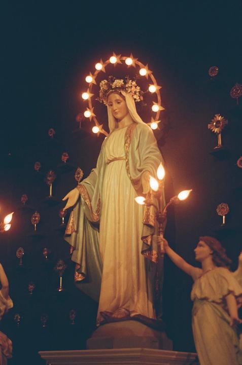 A statue of a woman with a halo of stars and flowers on her head stands in a dark room, with another statue of an angel holding a candelabra visible to the right.