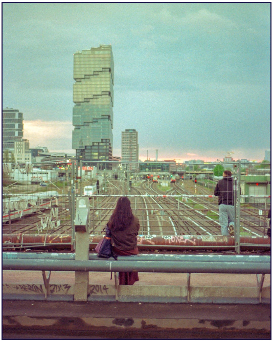 A woman and a man look out over train tracks and a city skyline at dusk.