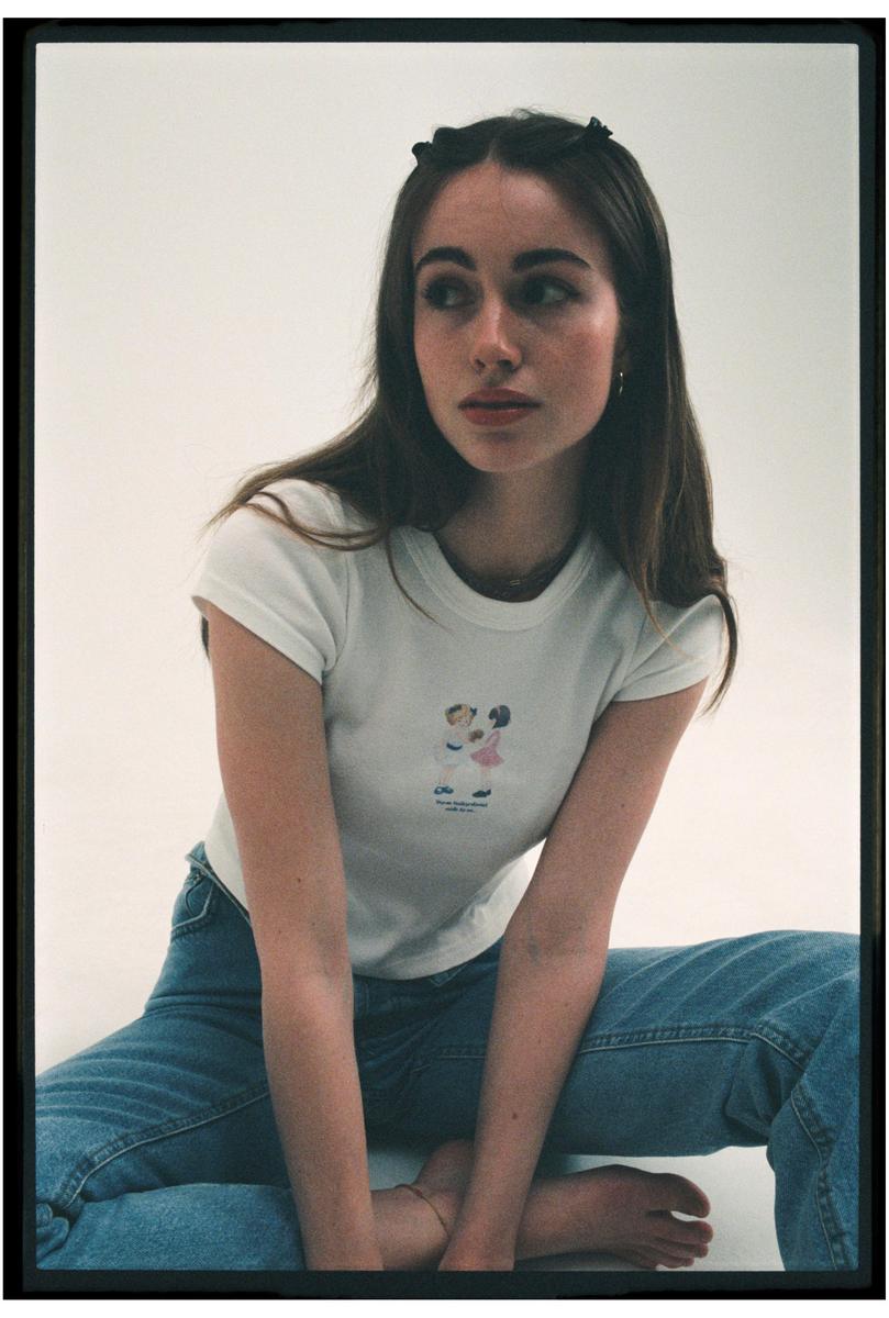 A young woman with long hair and hair clips sits on the floor, wearing a white t-shirt and jeans.