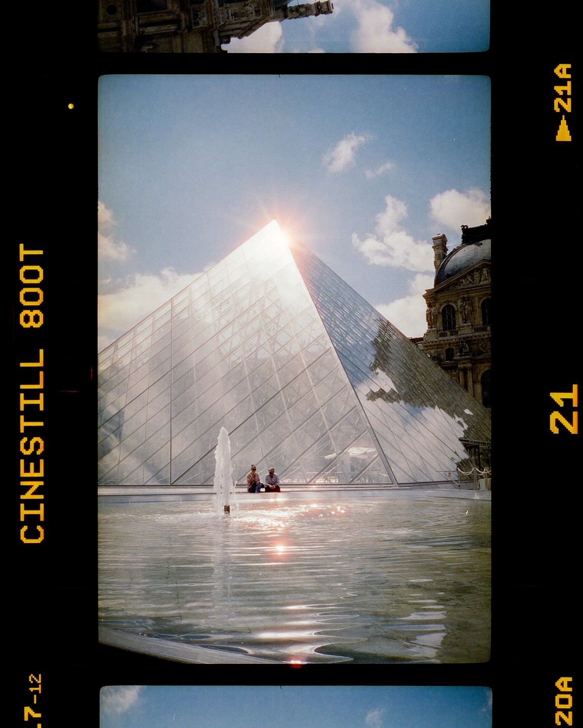 The glass pyramid of the Louvre is visible with a fountain in the foreground and two people sitting at its base.