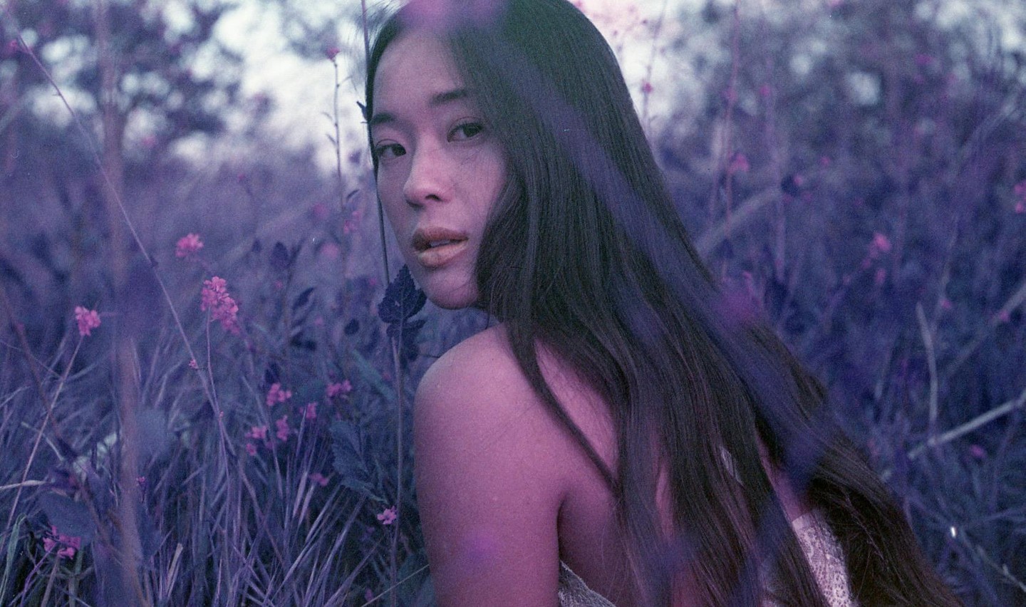 A woman with long dark hair looks over her shoulder while sitting in a field of purple flowers and grass.