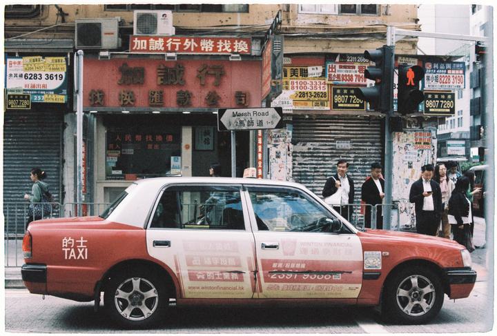 A red and white taxi is parked on a city street with people and buildings in the background.