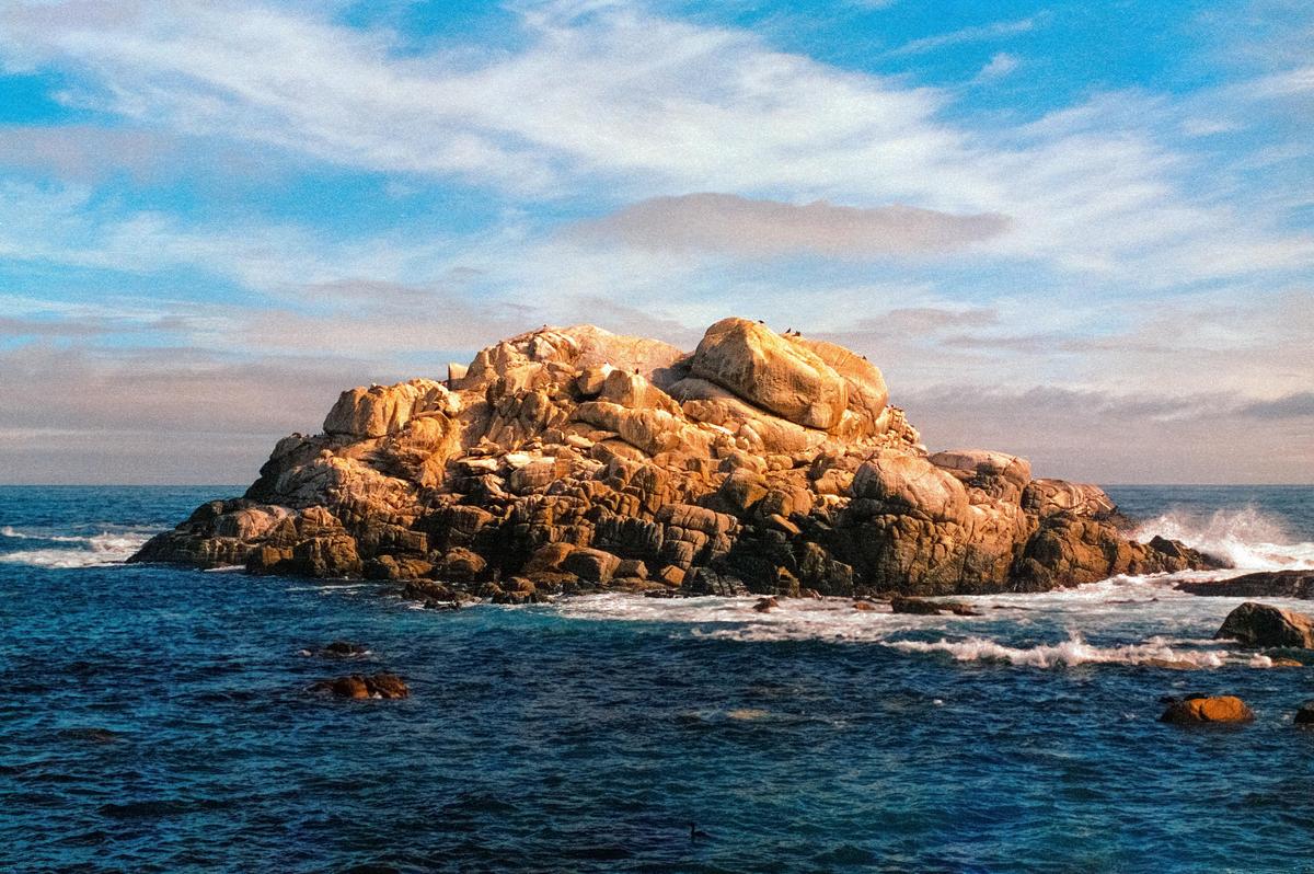 A large rock formation sits in the ocean under a blue sky with wispy clouds.