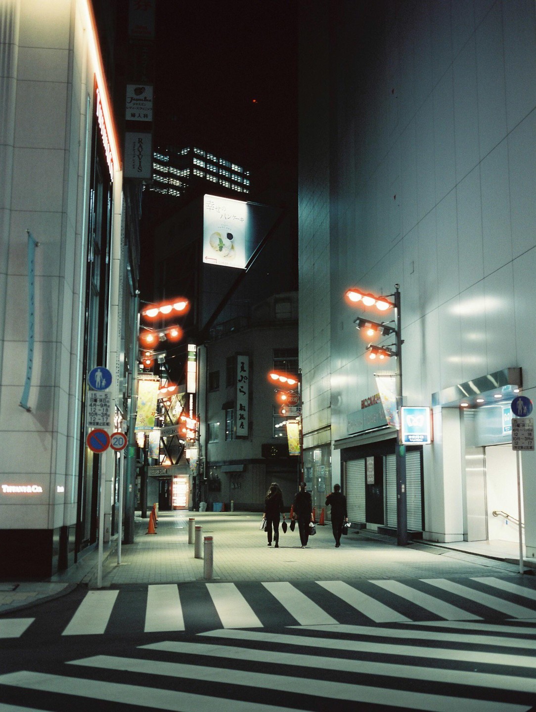 Three people walk away from the camera on a crosswalk at night in a city.