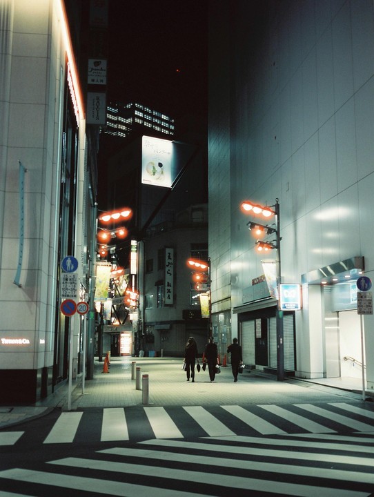 Three people walk away from the camera on a crosswalk at night in a city.