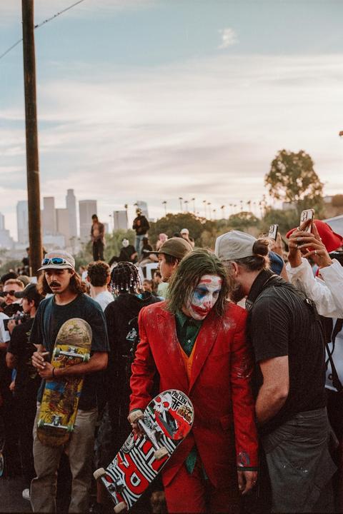 A person dressed as the Joker holds a skateboard while standing in a crowd of people on a street with a city skyline in the background.