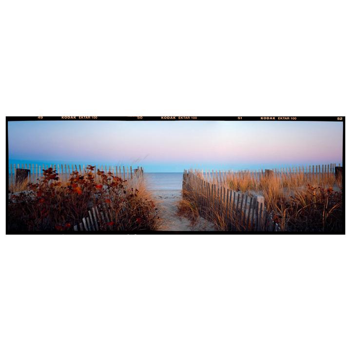 A sandy path leads through dunes and a wooden fence to a beach and ocean under a colorful sky.