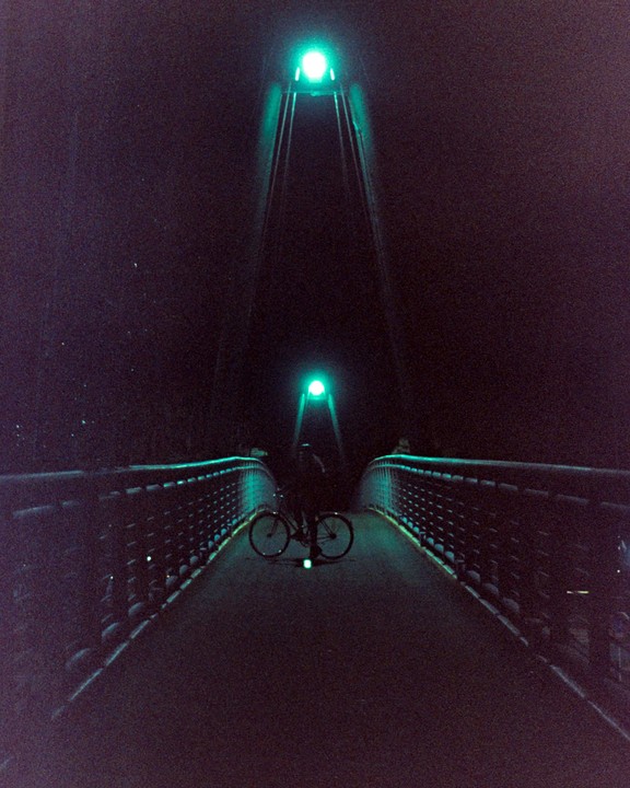 A person stands with a bicycle on a bridge illuminated by green lights at night.