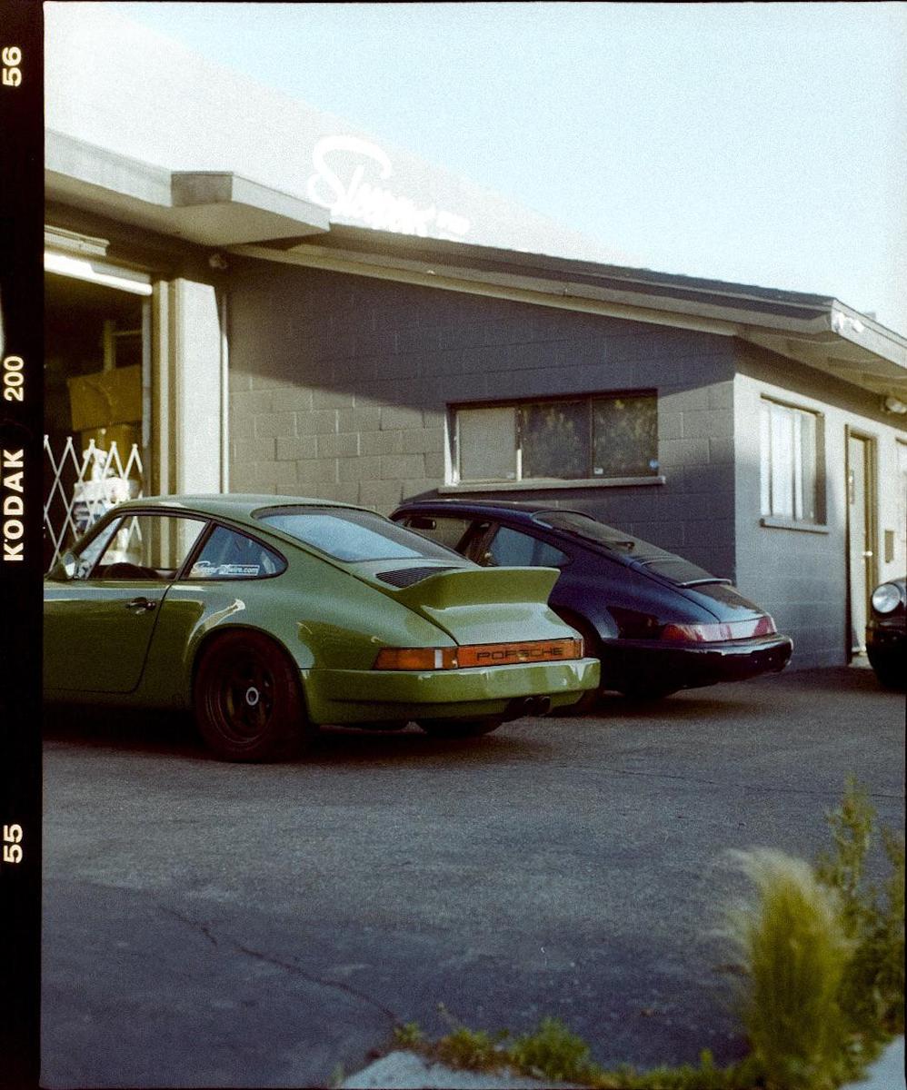 Two Porsche cars are parked outside a building with a light blue sky overhead.
