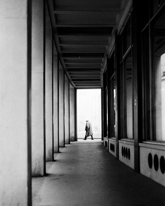 A person in a long coat walks down a long, narrow hallway with columns on one side and windows on the other.