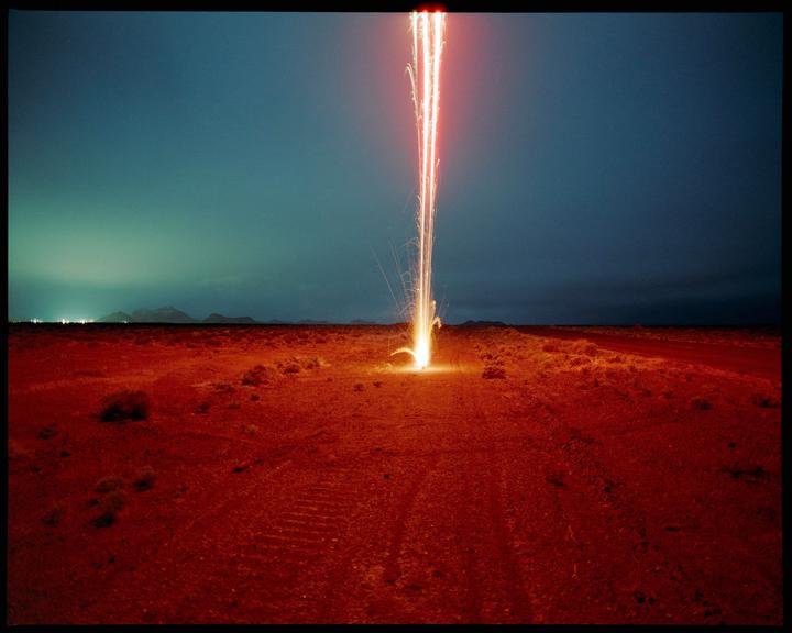 A bright red flare shoots into the dark blue sky from a red desert landscape.
