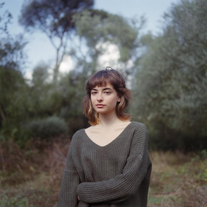 A woman with a bob haircut and bangs wears a green sweater and stands in a forest.