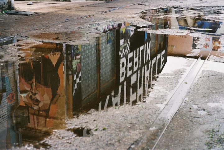A puddle on a street reflects a building with graffiti and the word "BERLIN" written on it.