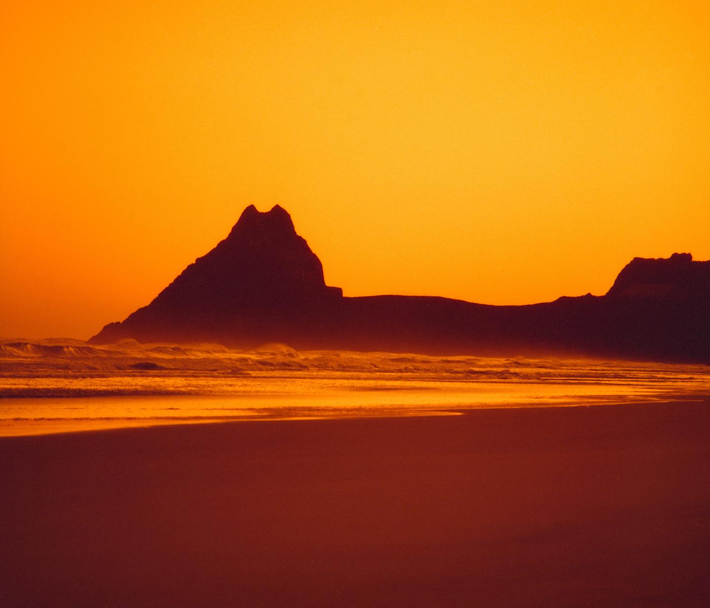 A silhouetted mountain range rises above a beach with waves under an orange sky.