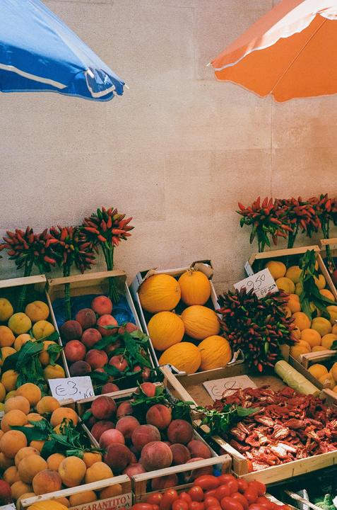 A market stall displays various fruits and vegetables in wooden crates under two umbrellas.