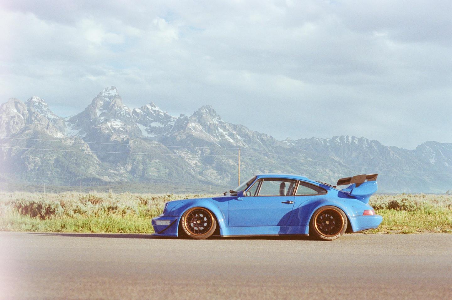 A blue sports car is parked on the side of a road with snow-capped mountains in the background.