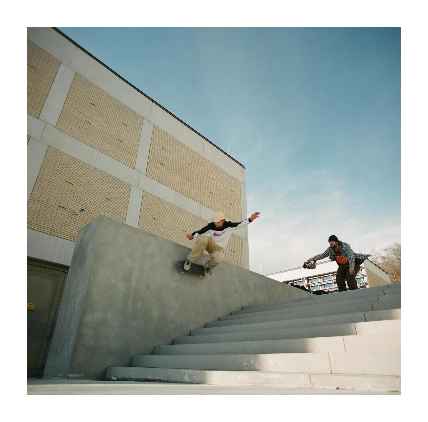A skateboarder grinds on a concrete ledge while another man films him on a sunny day.