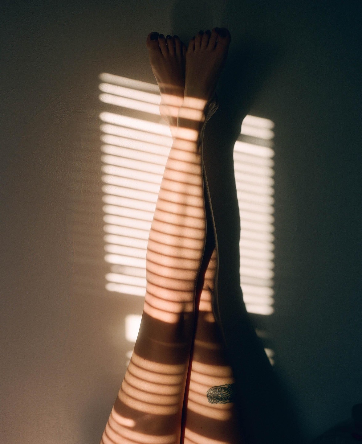 A person's legs are crossed with feet against a wall, illuminated by horizontal shadows from window blinds.