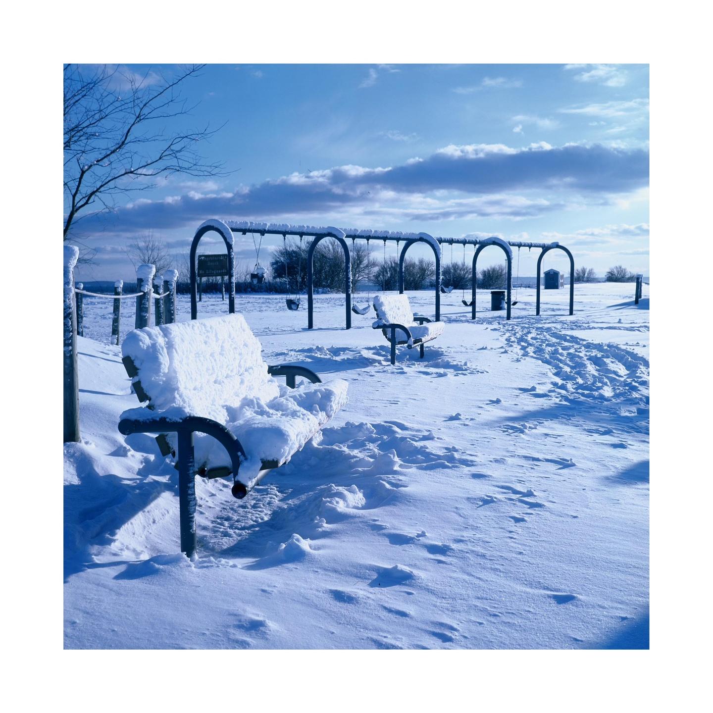 A snow-covered park bench sits in the foreground of a snowy playground with swings under a blue sky.