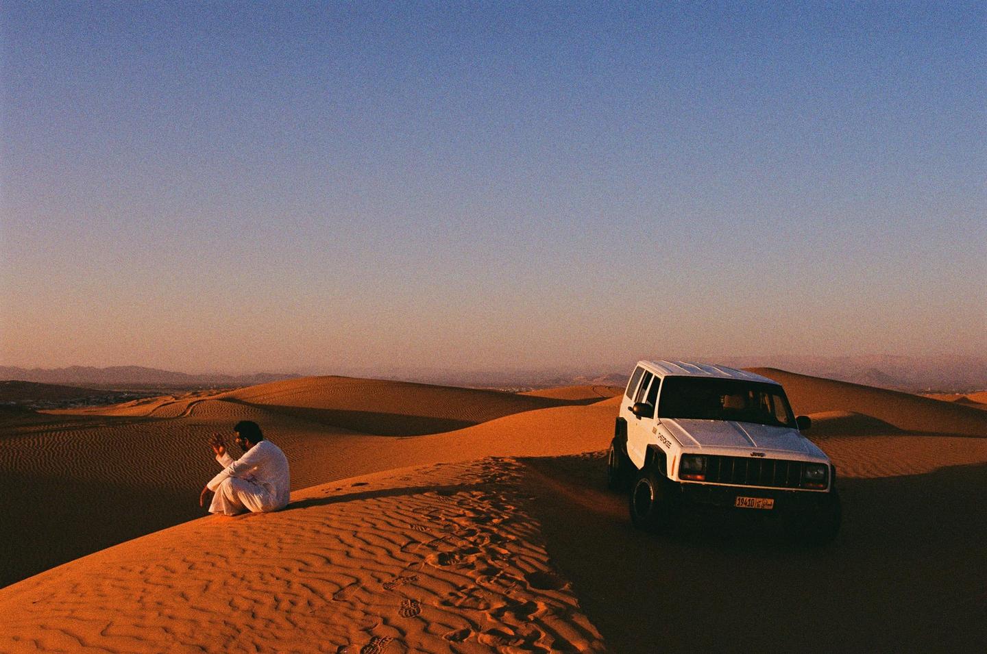 A man sits on a sand dune next to a white jeep at sunset.