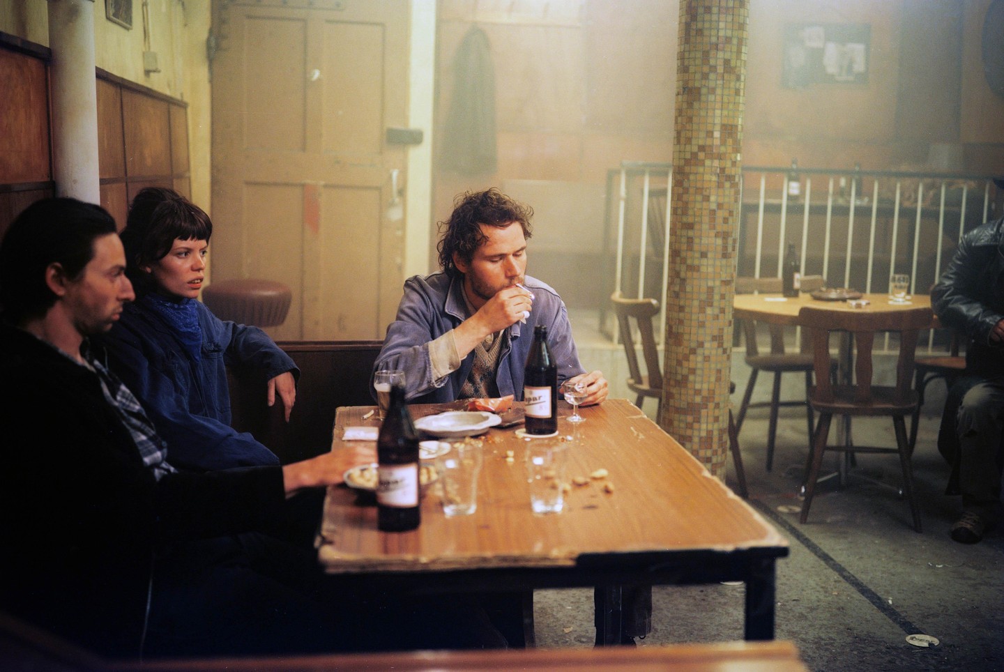 Three people sit at a wooden table in a hazy cafe, with one man smoking a cigarette.