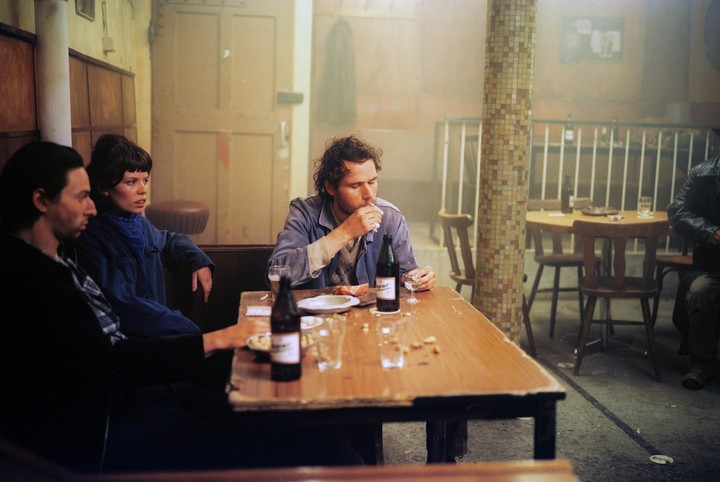 Three people sit at a wooden table in a hazy cafe, with one man smoking a cigarette.