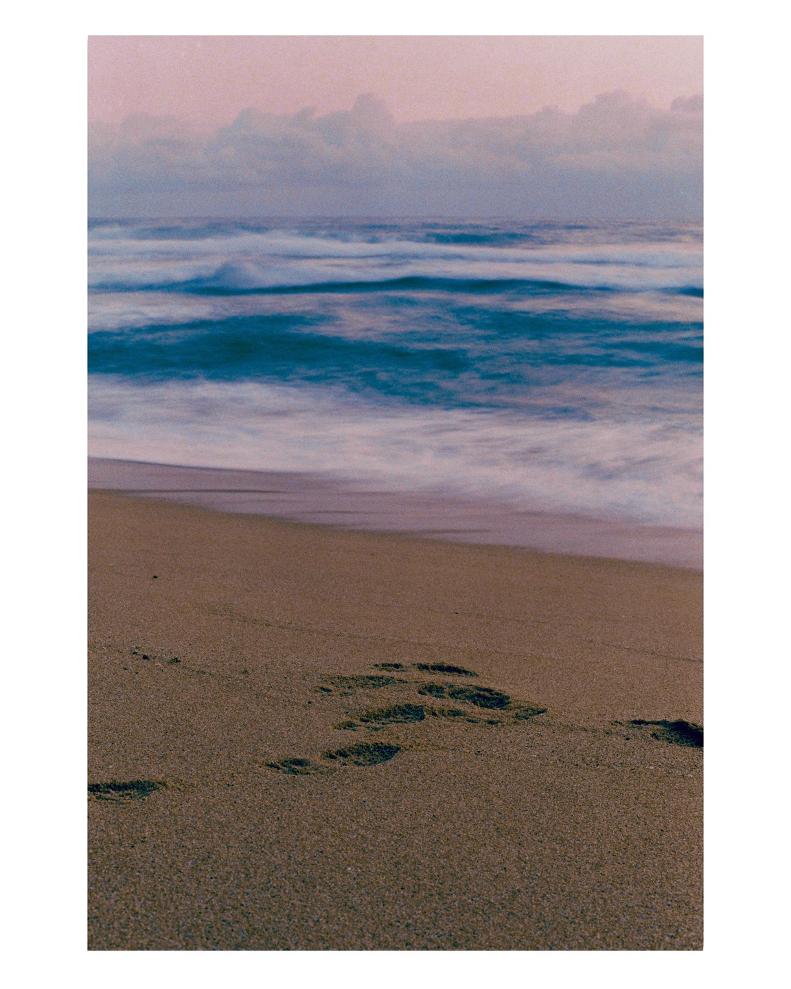 Footprints are visible in the sand next to the ocean under a cloudy sky.