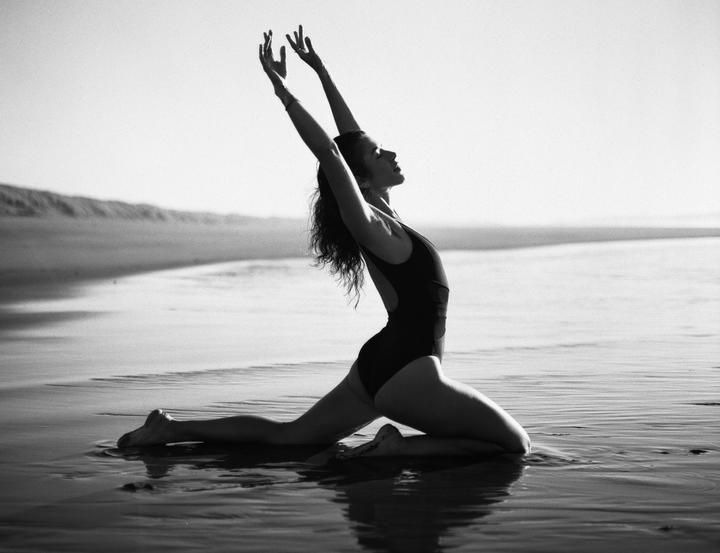 A woman in a swimsuit kneels in shallow water on a beach with her arms raised.