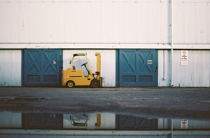 A yellow forklift is parked in front of a white corrugated metal building with two blue doors, and its reflection is visible in a puddle on the asphalt.