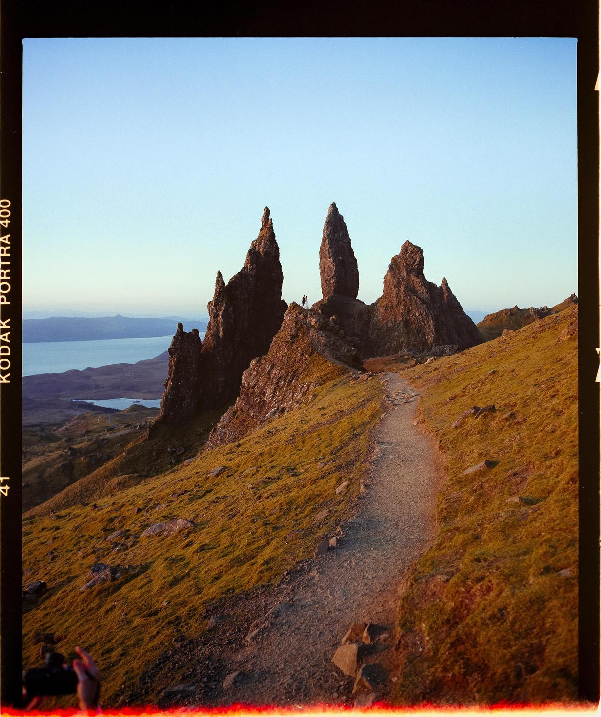 A narrow path leads up a grassy hill towards several large rock formations with a body of water and distant land visible in the background.