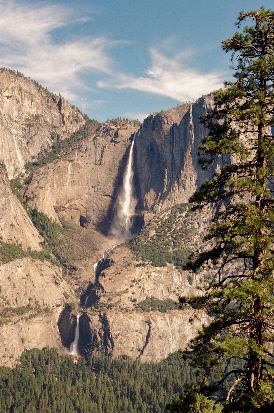 A tall waterfall cascades down a rocky mountain face, with a pine tree in the foreground.