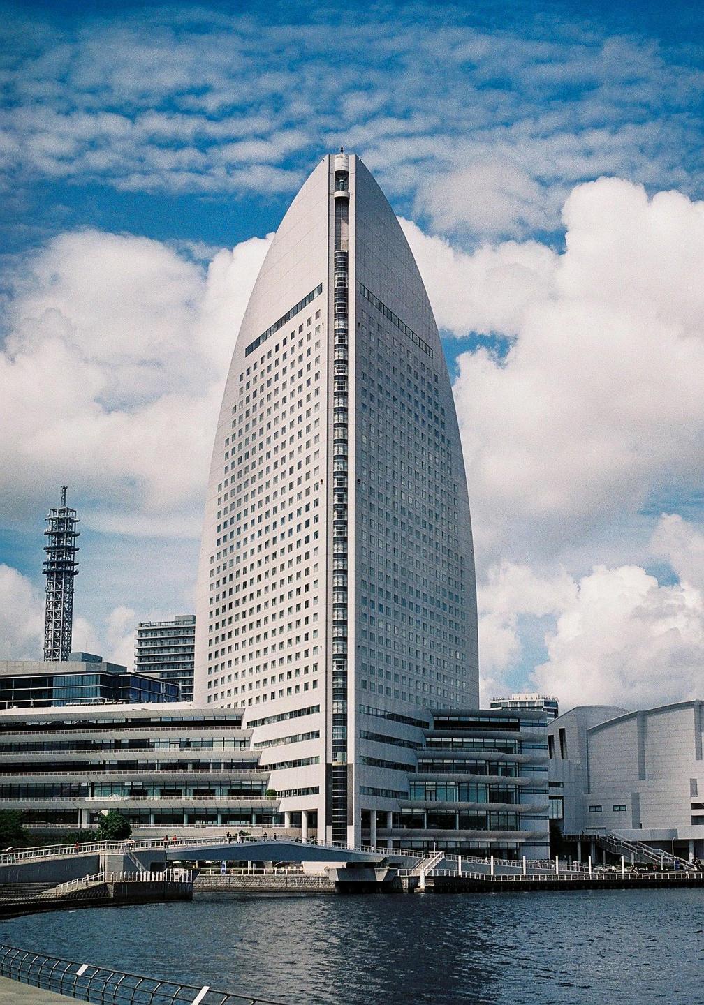 A tall, modern building with a curved facade stands beside a body of water under a cloudy sky.
