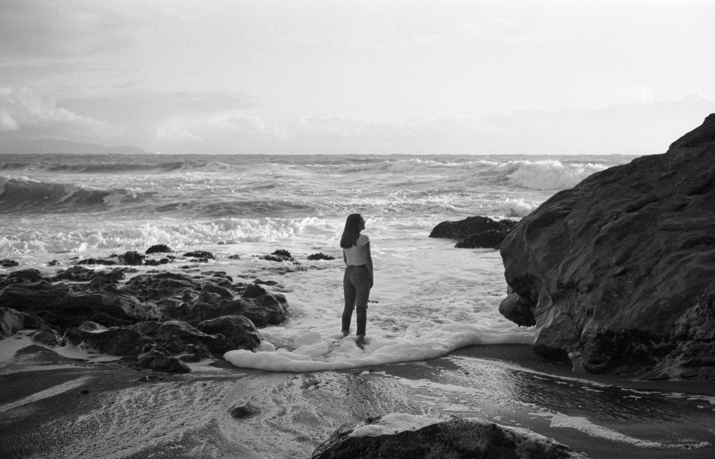 A woman stands in the foamy surf of a rocky beach, looking out at the ocean.