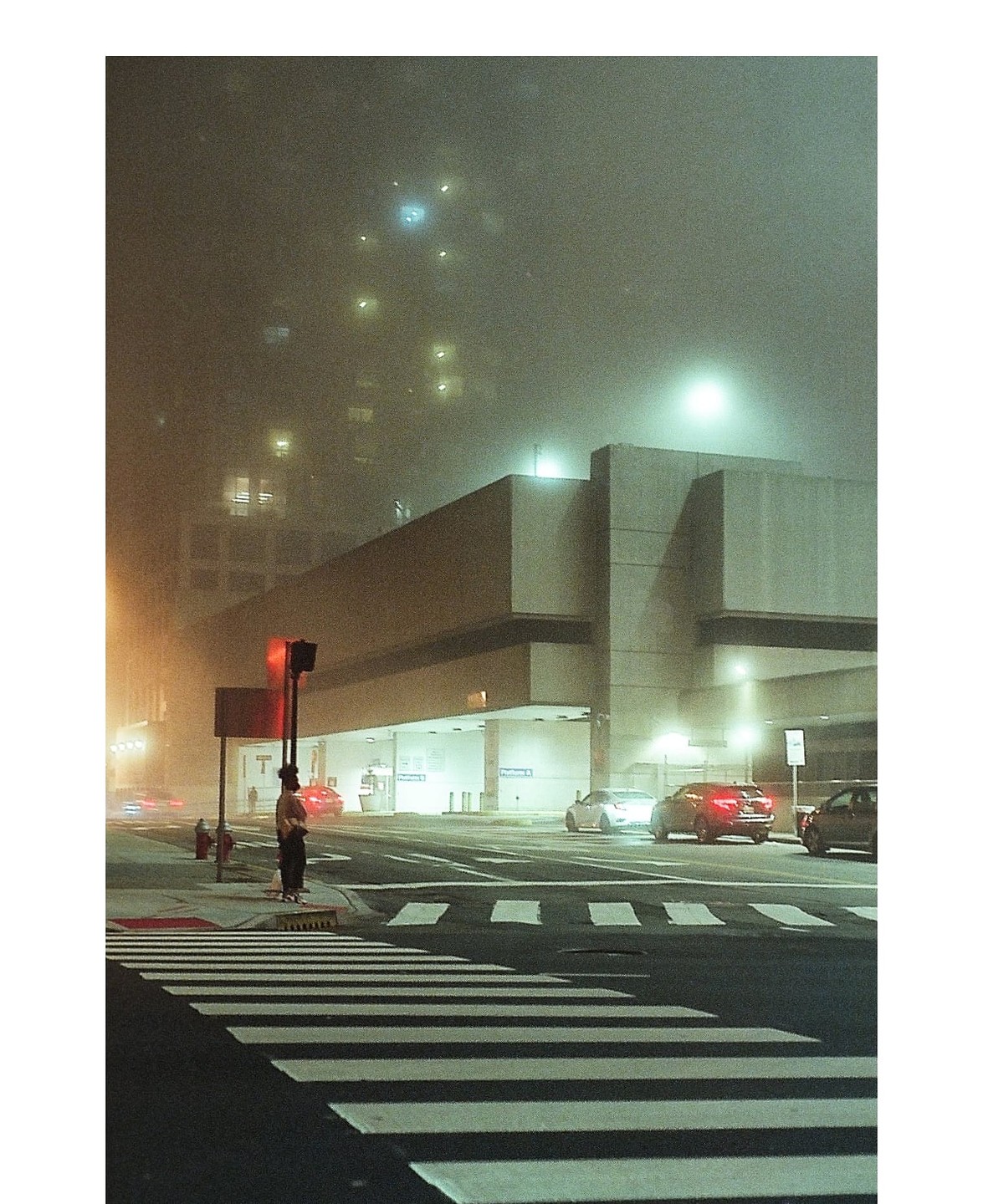 A person stands at a crosswalk on a foggy night with cars and buildings in the background.